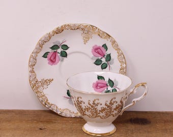 May include: A white porcelain teacup and saucer set featuring pink rose floral motifs and intricate gold filigree trim. The teacup has a pedestal base and a gold-accented handle, while the saucer displays matching pink roses and gold edges.