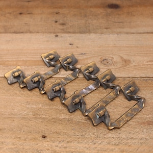 May include: Six antique brass drawer pulls are displayed. Each pull has a rectangular backplate, a twisted handle, and a square base. The brass has a weathered, aged look, with colour and texture variations. The pulls are arranged on a wooden surface.
