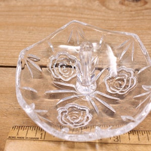 May include: A clear glass ring holder with a raised center post and a floral design. The holder has three roses etched into the glass.