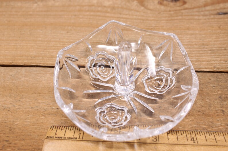 May include: A clear glass ring holder with a raised center post and a floral design. The holder has three roses etched into the glass.