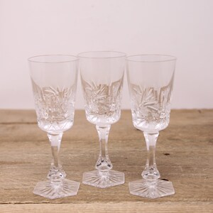 Peut inclure: Trois verres à vin en cristal transparent avec des motifs étoilés gravés. Chaque verre a une base hexagonale et une tige fine. Les verres sont disposés sur une surface en bois brun clair.
