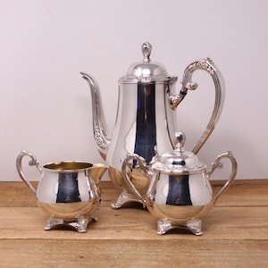 May include: A silver tea set, including a teapot, creamer, and sugar bowl. The teapot has a tall, curved spout and a decorative handle. The creamer and sugar bowl have ornate handles and feet. The set is displayed on a wooden surface.