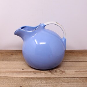May include: A pale blue ceramic jug with a rounded form and a curved spout. The handle is white with a floral pattern. The jug has a silver rim and is placed on a wooden surface. This vintage jug is suitable for serving drinks.