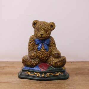 May include: A brown teddy bear doorstop with a blue bow sitting on a green, red, and blue patterned base.
