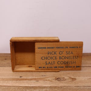 May include: A rectangular wooden box with a sliding lid. The front panel features blue text reading LAURENCE SWEENEY FISHERIES LTD. YARMOUTH, N.S. PICK O' SEA CHOICE BONELESS SALT CODFISH NET WT. 16 OZS. (ONE POUND) PRODUCT OF CANADA.