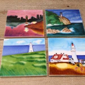 May include: Set of four ceramic tile coasters featuring colourful lighthouse scenes. Each coaster depicts a different lighthouse with a unique landscape, including a green grassy hill, a rocky coastline, and a blue ocean.