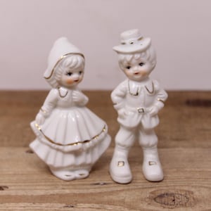 May include: A pair of white porcelain figurines of a boy and a girl. The boy is wearing a hat and a jacket, and the girl is wearing a dress and a bonnet. Both figurines have gold accents.