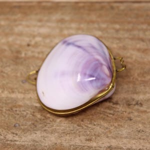 May include: A small, polished clam shell box with a smooth, light purple and white gradient surface. The shell is encased in a thin, gold-toned metal frame with a small hinge on one side and a decorative metal loop on the opposite side.