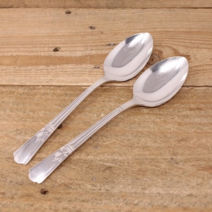 May include: Two silver spoons with ornate designs on the handles. The spoons are lying on a wooden surface.
