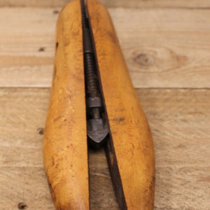 May include: A wooden shoe stretcher with a metal screw mechanism. The stretcher is made of light brown wood and has a slightly curved shape. The metal screw is visible in the center of the stretcher.