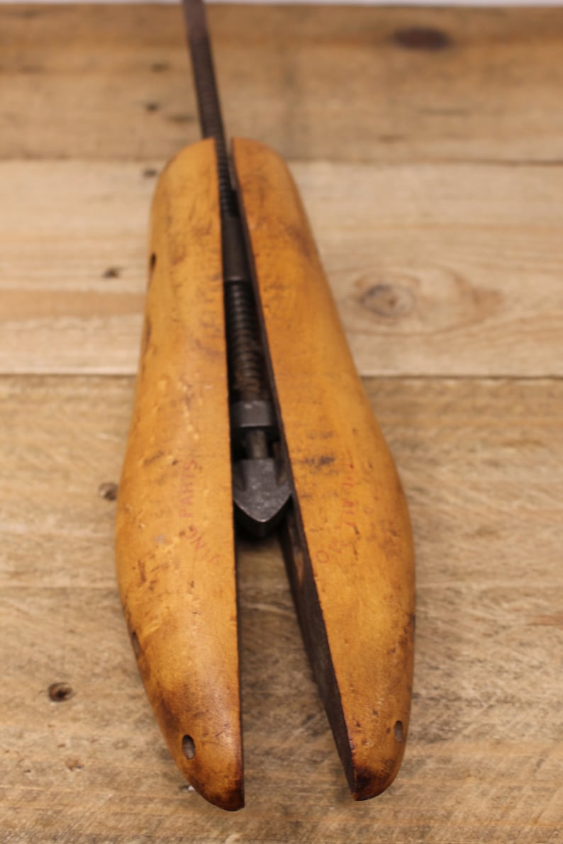 May include: A wooden shoe stretcher with a metal screw mechanism. The stretcher is made of light brown wood and has a slightly curved shape. The metal screw is visible in the center of the stretcher.