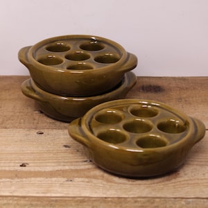 May include: Three green ceramic escargot dishes with six individual wells for serving snails. The dishes have two handles on the sides.