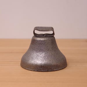 May include: A gray metal bell with a textured, weathered surface and a flat, rectangular loop handle at the top. The bell has a wide, flared base and a solid, cast appearance, showing a consistent metallic finish throughout its entire structure.