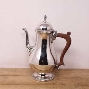 Peut inclure: Cafetière en argent avec une poignée en bois. La cafetière a un corps arrondi et un couvercle bombé avec un bouton.