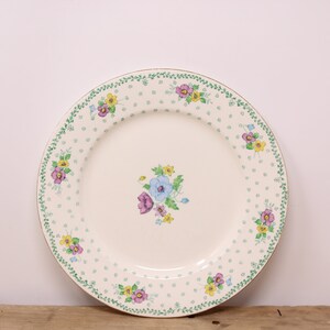 May include: A white ceramic plate with a green and gold floral border. The plate has a small floral design in the center and a white background with small green dots.