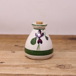 May include: A small, white ceramic bottle with a cork stopper. The bottle has a green band around the base and top, and a painted purple flower and green leaves design. The bottle is on a wooden surface.