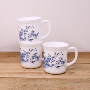 May include: Three white ceramic mugs with a blue floral design are stacked on a wooden surface. Each mug has a handle and a wavy rim. The floral pattern includes blue flowers and leaves.