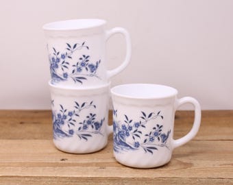 May include: Three white ceramic mugs with a blue floral design are stacked on a wooden surface. Each mug has a handle and a wavy rim. The floral pattern includes blue flowers and leaves.