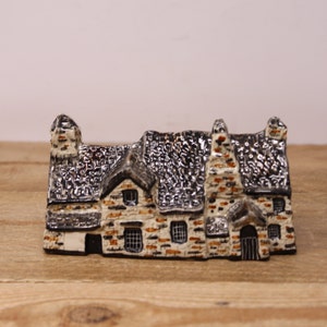 May include: A small ceramic model of a stone house with a black roof and white walls. The house has multiple windows and chimneys.