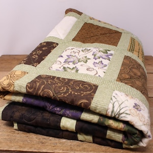 May include: Folded patchwork quilt in shades of brown, green, and cream. The quilt has a variety of patterns, including floral and geometric designs. The quilt is neatly folded, displaying the different fabrics and textures used in its construction.