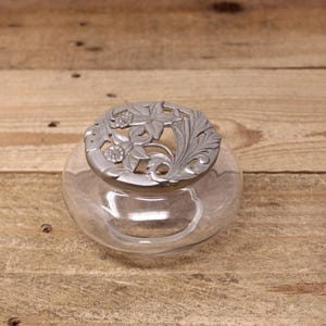 Peut inclure: Un pot en verre transparent avec un couvercle en argent orné d'un motif floral. Le couvercle présente un motif découpé de fleurs et de feuilles.
