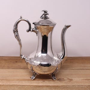 Peut inclure: Une cafetière argentée avec une anse et un bec verseur ornés. La cafetière présente un motif floral décoratif près de la base et une petite figurine d'oiseau sur le couvercle. Posée sur une surface en bois.