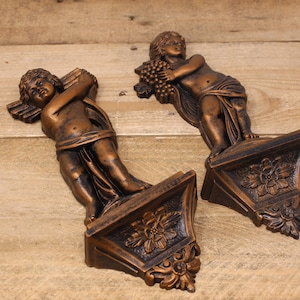 May include: Two ornate wall sconces featuring cherub figures. The cherubs are sculpted in a bronze-like finish and are holding a bunch of grapes and a scroll. The sconces have a decorative floral design.