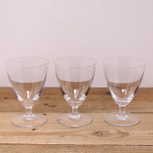 May include: Three clear glass wine glasses with a stemmed base. The glasses are empty and arranged in a row on a wooden surface.