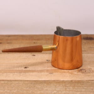 May include: A copper-colored, small saucepan with a wooden handle and brass fitting. The interior of the pan is a silver-gray color. The handle is a light brown color. The saucepan is sitting on a wooden surface.