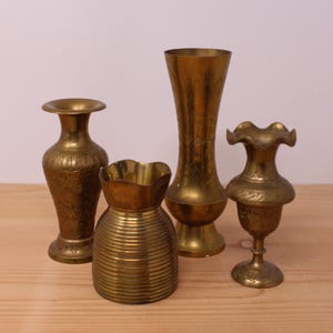 Peut inclure: Un ensemble de quatre vases en laiton de formes et de tailles variées. Trois vases présentent des motifs floraux gravés complexes et des bords évasés ou ondulés, tandis qu'un vase plus court et large affiche une texture nervurée horizontale.