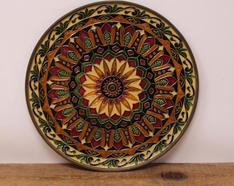 May include: A decorative, round plate with an intricate, symmetrical design. The plate features a central starburst pattern in yellow, surrounded by red, green, and brown geometric shapes. The outer edge has a green leaf pattern and a metallic rim.
