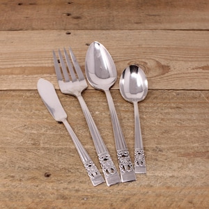 May include: Four pieces of cutlery, a butter knife, fork, and two spoons, all with ornate floral designs on the handles. The cutlery is silver in colour and is laid out on a wooden surface.