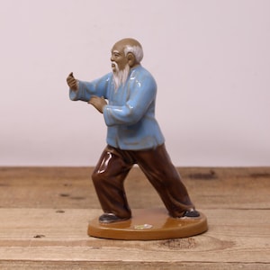 Peut inclure: Figurine en céramique d'un vieil homme en position de Tai Chi. La figurine porte une veste bleu clair et un pantalon marron. La figurine est sur une base marron et a une barbe blanche.