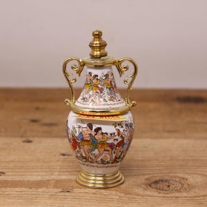 May include: A decorative ceramic perfume bottle with a gold-colored lid and handles. The bottle features a painted scene of figures in classical attire. The base and lid are trimmed with gold. The text "NINA RICCI" is visible on the bottle.