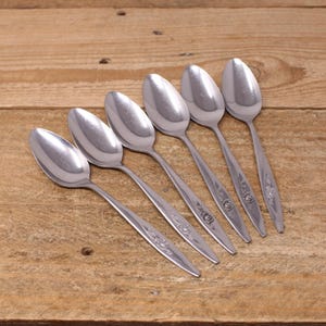 May include: Six stainless steel spoons arranged diagonally on a wooden surface. The spoons have a classic design with elongated bowls and floral detailing on the handles. The spoons are reflective, showing the light.