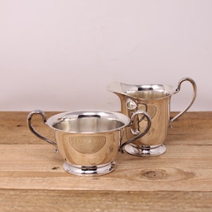 May include: A silver creamer jug and sugar bowl set. The creamer has a curved handle and spout. The sugar bowl has two curved handles. Both pieces have a pedestal base and are displayed on a wooden surface.