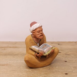 May include: A ceramic figurine of a seated man reading a book. The figure wears a turban and a long robe in shades of yellow and brown. The book has colorful pages and is held in the man's lap. The figurine is set against a light background.