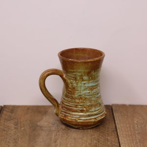 May include: Handmade ceramic mug with a brown rim and handle, and a textured, turquoise and brown body. The mug has a cylindrical shape and is suitable for hot or cold beverages. The mug is displayed on a wooden surface.