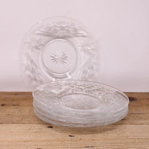 May include: A stack of clear glass plates featuring a starburst design in the centre and a diamond pattern around the edges. One plate is tilted behind the stack. The plates are on a wooden surface.