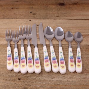 May include: A set of stainless steel silverware with white handles decorated with a floral pattern in yellow, pink, and blue. The set includes forks, knives, and spoons, arranged on a wooden surface.