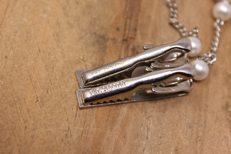May include: Close-up of two silver metal clips with the text "PAT. 2435761" engraved on each. The clips are attached to a pearl necklace.