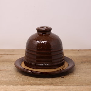 May include: A brown ceramic butter dish with a domed lid and a matching plate. The dish has a glossy finish and horizontal ridges. The plate is a lighter brown color and has a raised edge. The butter dish is sitting on a wooden surface.