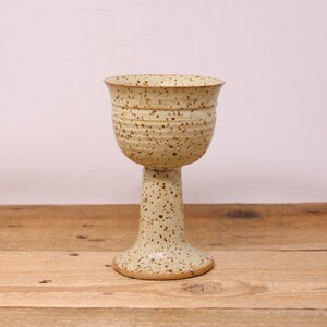 May include: A handmade ceramic goblet with a speckled, light beige finish. The goblet features a wide, rounded bowl and a tall, slender stem, resting on a flared base. The piece is approximately 15 cm tall.