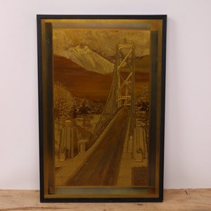 May include: A framed gold-toned embossed image of a bridge with a mountain range in the background. The bridge is a suspension bridge with a road leading to it. The image is framed in a black frame with a gold-toned border.