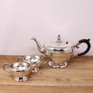 May include: A silver teapot and two creamer pitchers with black handles. The teapot has a round body and a lid with a knob. The creamers have a round body and a spout. The set is on a wooden surface.