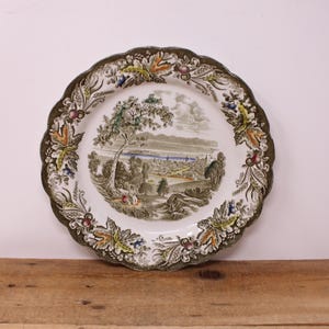 May include: A round ceramic plate with a scalloped edge featuring a central landscape illustration of a city near a body of water with trees and a figure. The border is decorated with a detailed pattern of green leaves, wheat, and small fruits.
