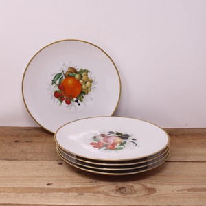 May include: White ceramic plates with gold rims. One plate features a vibrant orange, grapes, and berries design. A stack of plates with a floral and fruit design is visible. The plates are on a wooden surface.