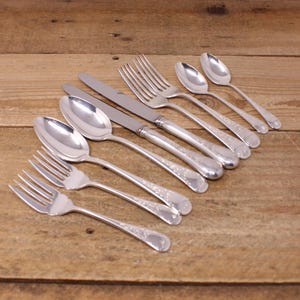 May include: An assortment of polished silverware, including forks, spoons, and knives, arranged on a wooden surface. The silverware has a decorative pattern on the handles and reflects the light. The arrangement suggests a formal dining setting.