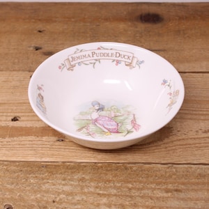 May include: A white ceramic bowl featuring a central illustration of Jemima Puddle-Duck in a bonnet and shawl, surrounded by green grass and pink flowers. The rim displays a banner with the text Jemima Puddle-Duck and small floral accents.