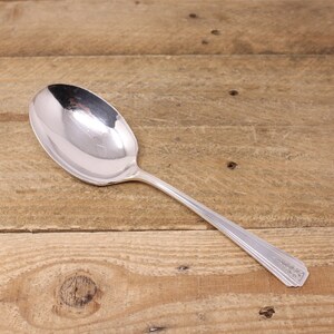 May include: A polished silver spoon with a large, oval bowl and a long, slender handle. The handle features a decorative pattern near the end. The spoon is placed on a wooden surface with a natural grain.